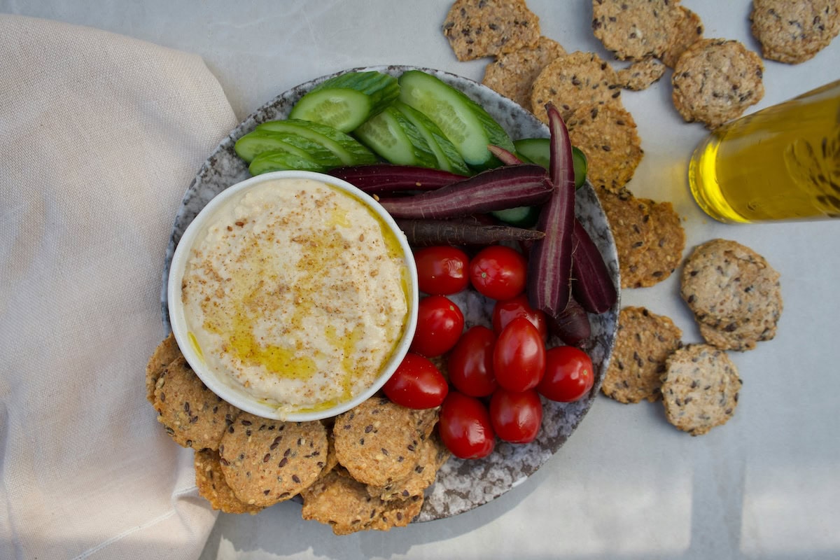 A top-down view of a hummus platter with cucumber slices, cherry tomatoes, purple vegetable sticks, and whole-grain crackers, styled on a grey surface